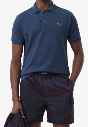 Mann trägt ein marineblaues Poloshirt mit grünem Krokodil-Logo und dunkel gemusterte Shorts, hält eine passende marineblaue Jacke in der Hand.