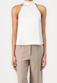 Femme portant un haut dos nu blanc sans manches avec un col haut et un pantalon beige ajusté, se tenant devant un fond uni.