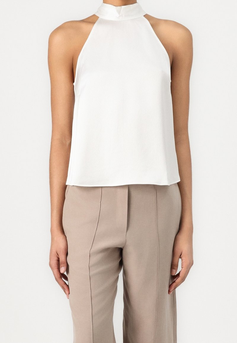 Femme portant un haut dos nu blanc sans manches avec un col haut et un pantalon beige ajusté, se tenant devant un fond uni.