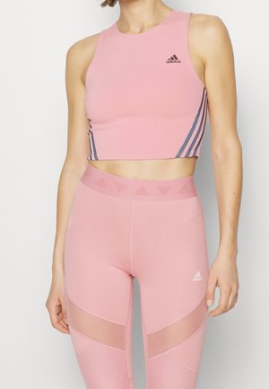 Kobieta w różowym bezrękawniku Adidas crop top i legginsach z wysokim stanem, z panelami siateczkowymi i detalami logo na białym tle.
