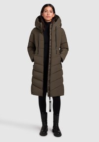 Abrigo largo de plumas en verde oliva con capucha, que cuenta con cierre de cremallera, bolsillos laterales y un diseño acolchado. Llevado sobre un conjunto negro y botas negras.