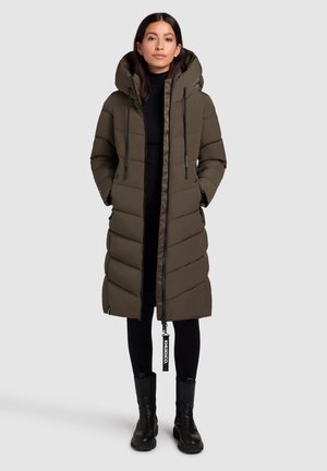 Abrigo largo de plumas en verde oliva con capucha, que cuenta con cierre de cremallera, bolsillos laterales y un diseño acolchado. Llevado sobre un conjunto negro y botas negras.