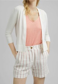 Kevyt valkoinen neuletakki vaaleanpunaisen V-kaula-aukkopaidan päällä, yhdistettynä valkoisiin shortseihin, joissa on punaiset raidat ja puunapit vyötäröllä.