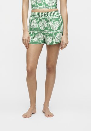 Γυναίκα με πράσινες και λευκές σορτς με σχέδιο κοχυλιών και ταιριαστό κοντό τοπ, ξυπόλητη μπροστά σε μονόχρωμο φόντο.