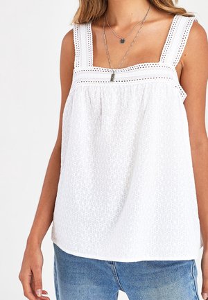 Hvid ærmeløs øjelynlace top med firkantet halsudskæring og brede stropper, båret med lagdelte sølvsmykker og blå jeans.