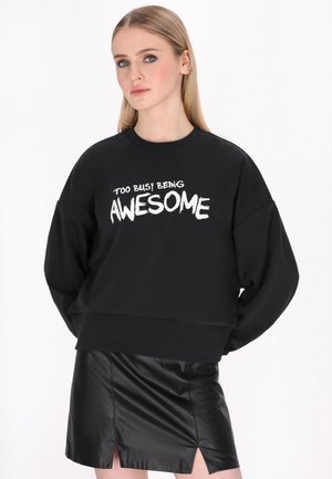 Młoda kobieta o prostych blond włosach nosi czarną bluzę z napisem „Too busy being awesome” oraz czarną skórzaną spódnicę.