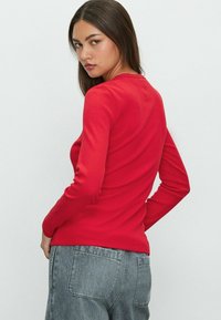 Vrouw met lang donker haar, die een strakke rode lange mouwen top en grijze jeans draagt, met een lichte draai en handen in de achterzakken.