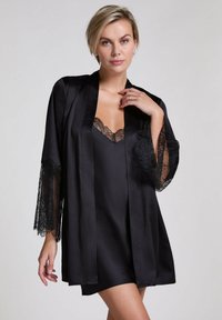 Accappatoio in raso nero con maniche rifinite in pizzo, caratterizzato da un profondo scollo a V sopra un vestito sottile nero coordinato. Texture liscia e design elegante.