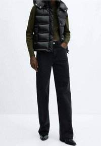 Czarna pikowana kamizelka z kapturem o błyszczącej fakturze, zestawiona z ciemnymi jeansami o prostych nogawkach i czarnymi butami z ostrym noskiem.