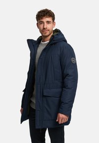 Marineblaue Steppjacke mit Kapuze, zwei vorderen Taschen und einem aufgenähten Logo am linken Ärmel. Strukturierter Stoff mit Reißverschluss.