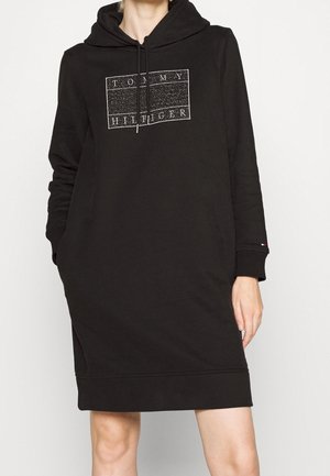 Femme portant une robe noire à manches longues avec capuche et logo "Tommy Hilfiger" sur la poitrine ainsi que des poches latérales, debout devant un fond uni.