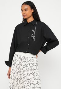 Γυναίκα με μαύρο πουκάμισο κουμπωτό, κεντημένο με τη λέξη "Karl", και λευκή πλισέ φούστα με μοτίβο μαύρης χειρόγραφης γραφής.