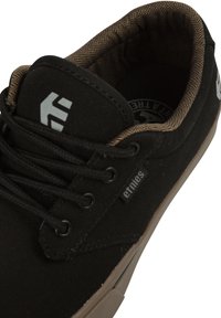 Black canvas sneaker with brown herringbone lining, featuring six eyelets, black laces, and a prominent grey logo detail on the side.
