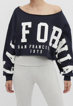 Marineblaues kurzes Sweatshirt mit dem Text "California San Francisco 1975", schulterfrei getragen, kombiniert mit hellgrauen, hoch taillierten Leggings.