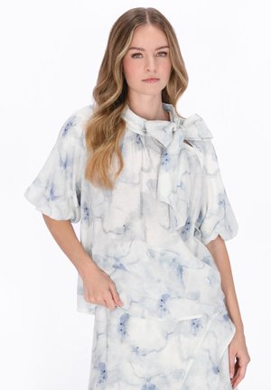 Kobieta o falowanych włosach, ubrana w jasną bluzkę i spódnicę z wzorami w odcieniach niebiesko-szarych przypominającymi akwarele oraz dużą kokardą przy dekolcie.