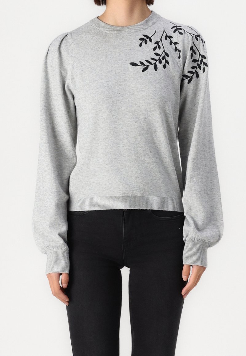 Grå sweater med lange ærmer, rund halsudskæring og puffet skulderdetalje. Har et sort broderet blade-design på venstre skulder.