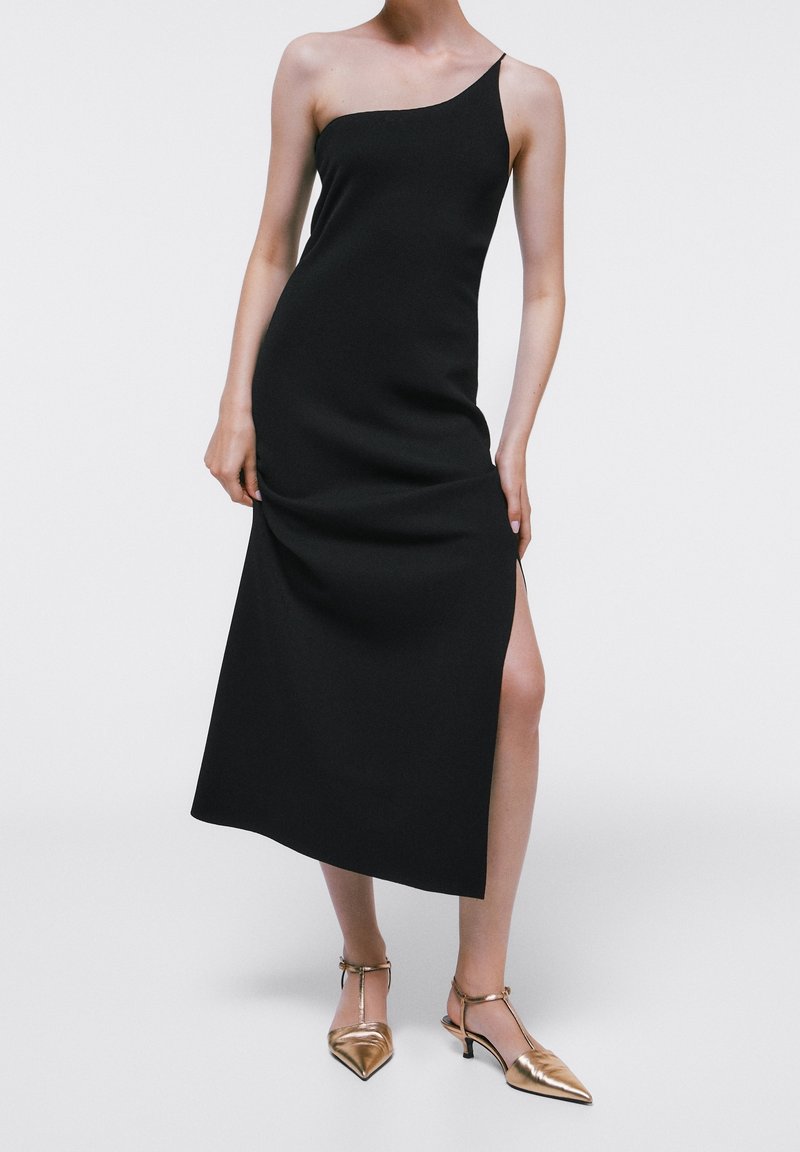 Femme portant une robe midi noire à une épaule avec une fente haute sur la cuisse et des chaussures pointues dorées métalliques à talon bas.