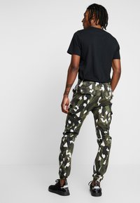 Fekete póló, amelyet terepmintás cargo nadrággal párosítottak, sötétzöld és fehér geometrikus minták keverékével. Slim-fit szabás, rugalmas mandzsettákkal.