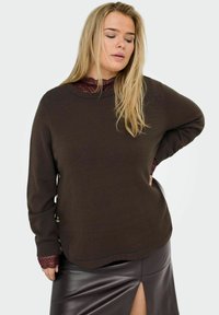 Femme blonde portant un pull brun foncé avec des boutons sur le côté et une jupe en cuir noire avec une fente à l'avant.