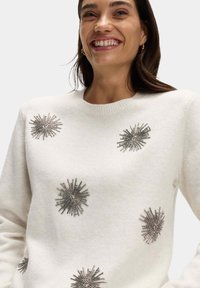 Bílý svetr s texturovanými, metalickými ozdobami připomínajícími tryskající hvězdy, s kulatým výstřihem a dlouhými rukávy. Měkká tkanina.