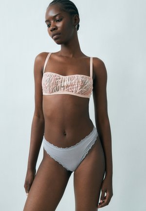Femme aux cheveux tressés portant un soutien-gorge rose clair à motifs et une culotte gris clair bordée de dentelle, debout devant un fond uni.