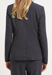 Veste de costume bleu marine foncé avec des rayures blanches verticales, coupe structurée et une fente à l'arrière. Le matériau semble texturé avec un design sur mesure.