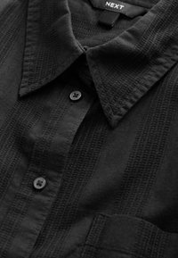 Chemise noire à col avec un motif rayé texturé, fermeture à boutons noirs à l'avant, et une poche poitrine unique.