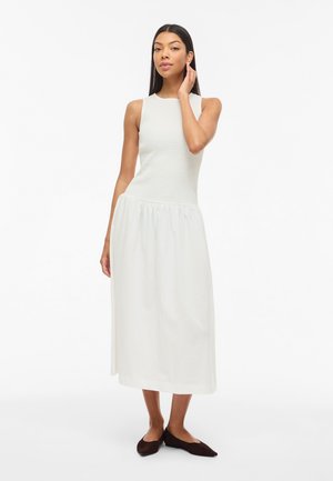 Γυναίκα με λευκό αμάνικο φόρεμα μήκους μέχρι τη μέση της γάμπας και μαύρα φλατ παπούτσια στέκεται με το ένα χέρι να αγγίζει τα μαλλιά της, μπροστά σε ένα απλό λευκό φόντο.