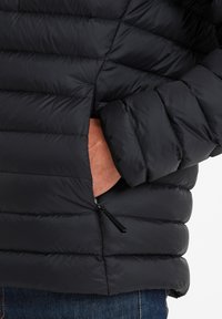 Black puffer jacket featuring a quilted design, soft texture, and zippered side pocket, worn over denim blue jeans.