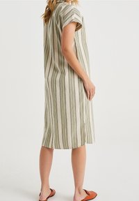 Femme portant une robe à manches courtes, arrivant au genou, avec des rayures verticales vertes et beiges, associée à des chaussures brunes à enfiler.