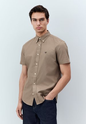 Fiatal férfi, kezét zsebében tartva, bézs színű rövid ujjú gombos inget és sötét farmert visel, sima világos háttér előtt.