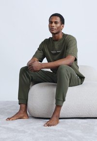 Πράσινο βαμβακερό μπλουζάκι και παντελόνι σε χαλαρή γραμμή, με λογότυπο στο στήθος. Η φιγούρα κάθεται σε ένα ανοιχτό, υφασμάτινο πουφ.