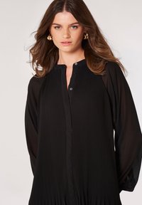 Schwarzes, plissiertes Kleid mit rundem Ausschnitt, langen Ärmeln, Knopfverschlüssen und einem fließenden Schnitt aus strukturiertem Stoff.