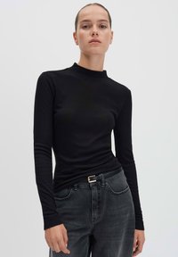 Vrouw in een strak zwart langekleurige top en vervaagde zwarte spijkerbroeken met een zwarte riem, staand tegen een effen lichte achtergrond.