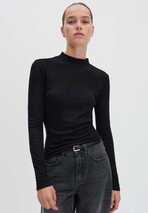 Vrouw in een strak zwart langekleurige top en vervaagde zwarte spijkerbroeken met een zwarte riem, staand tegen een effen lichte achtergrond.