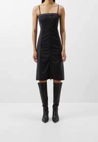 Sort ærmeløs kjole med lynlås foran, struktureret bodice og lige silhuet, parret med knæhøje sorte hæle støvler.