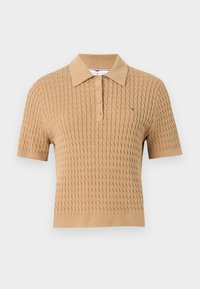 Beżowa koszulka polo z krótkim rękawem, pleciona w kabel, z plisą na guziki i małym logo w kolorach czerwonym, białym i niebieskim po lewej stronie klatki piersiowej.