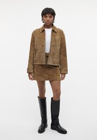 Giacca e gonna in suede marrone con decorazioni metalliche. La modella indossa una maglietta bianca e stivali neri al ginocchio. Texture liscia, chiusura con zip.