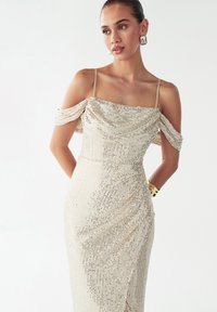 Obleka z biseri v svetlo zlati barvi z brezzračnimi naramnicami, nabrano korzetno zgornjo del in oprijetim krilom z navpičnimi črtami in razpršenimi svetlečimi palčkami.