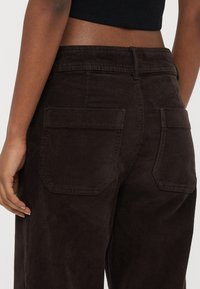 Calças de bombazina castanhas com cintura ajustada, apresentando dois bolsos traseiros e textura canelada vertical. O tecido parece macio e resistente.