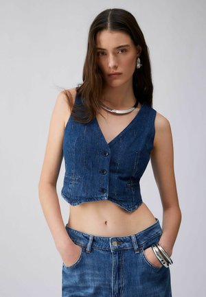 Kobieta ubrana w niebieską jeansową kamizelkę bez rękawów i jeansy, z rękami w kieszeniach, ozdobiona srebrną biżuterią, w tym naszyjnikiem, kolczykami i bransoletkami.