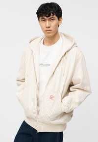 Νεαρός άνδρας που φοράει κρεμ φούτερ με φερμουάρ και λεπτές ρίγες πάνω από λευκό μπλουζάκι Hollister και σκούρο τζιν, στέκεται με τα χέρια στις τσέπες.