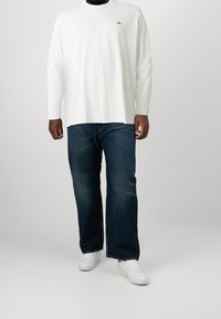 Man som bär en vit långärmad tröja med en liten grön logotyp, mörkblå jeans och vita sneakers mot en enkel ljus bakgrund.