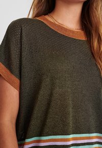 Frau trägt ein dunkelolivfarbenes Stricktop mit orangefarbenen Akzenten an den Ärmeln und am Kragen, das über perforierte Schulterdetails und einen gestreiften Bund an der Taille verfügt.