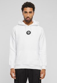 Felpa bianca con tasca a marsupio, cappuccio regolabile con lacci e un logo nero circolare sul petto. Realizzata in tessuto morbido e texture.