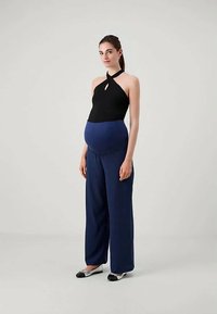 Gravid kvinne som står, iført en svart halterneck-top og løse mørkeblå mammabukser med sølv- og svarte leppestift, foran en ensfarget bakgrunn.