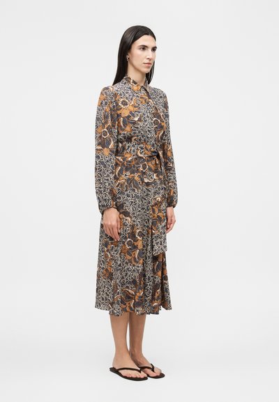 Γυναίκα με μακριά σκούρα μαλλιά, φορώντας ένα μίντι φόρεμα με μακριά μανίκια και φλοράλ σχέδιο και μαύρα σαγιονάρα, στέκεται μπροστά σε απλό φόντο.