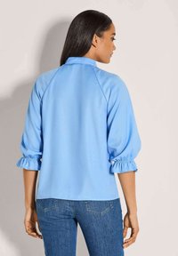 Vrouw met lang, donker haar, gekleed in een lichtblauwe blouse met driekwartmouwen en gerimpelde manchetten, gecombineerd met blauwe spijkerbroeken, met haar rug naar de camera gekeerd.