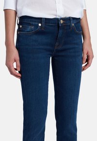Jeans en denim bleu foncé avec une coupe slim, cinq poches et des coutures contrastées. Présente une fermeture à bouton et des accents en métal.
