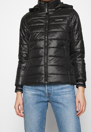 Schwarze, gesteppte Calvin Klein Jacke mit Frontreißverschluss, Seitentaschen und Kapuze, getragen zu hellblauen Jeans, Hände entspannt an den Seiten.